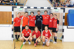 Neun Personen in roten und schwarzen Sporttrikots stehen und knien vor einem Hallentor.