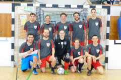 Acht Personen in Sportkleidung vor einem Handballtor, eine Person in schwarzer Torwartkleidung, Ball am Boden.