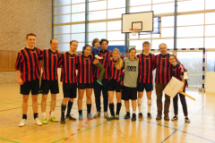 Gruppe von zehn Personen in rot-schwarzen Trikots und Sportkleidung in einer Sporthalle, Basketballkorb im Hintergrund.