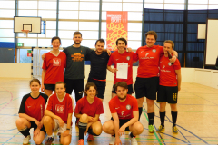 Acht Personen in Sportkleidung stehen und knien in einer Turnhalle; zwei Basketballkörbe und ein Banner im Hintergrund.