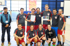 Gruppe von zehn Männern in Sportkleidung in einer Halle; zwei halten Papiere, links steht eine weitere Person.
