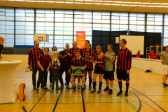 Gruppe von Personen in Sportkleidung in einer Sporthalle, einige halten einen Pokal und eine Kiste.