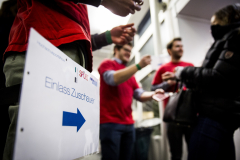 Schild mit Aufschrift „Einlass Zuschauer“ und blauem Pfeil; daneben mehrere Personen in roten Shirts und eine Person in schwarzer Jacke.