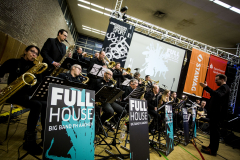 Große Big Band mit Saxofonen, Trompeten und Posaunen auf einer Bühne; vorne Schilder mit „FULL HOUSE“.