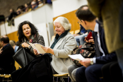 Mehrere sitzende Personen in einem Saal, einige halten Papierblätter; im Hintergrund weitere Zuschauer.