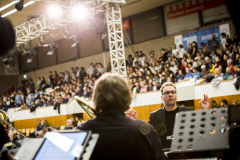 Orchesterprobe in einer Halle mit Publikum auf den Tribünen und beleuchteter Bühne.