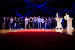Personengruppe auf einer Bühne mit blauem Licht, rotem Teppich und zwei Stehtischen rechts.