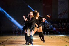 Drei Tänzerinnen in schwarzen Outfits auf einer Sporthalle-Bühne, mit Lichtstrahlen und Publikum im Hintergrund.