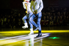 Zwei Personen in Cowboyhüten tanzen auf einer beleuchteten Bühne in einer Halle.