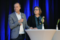 Zwei Personen stehen hinter einem weißen Stehpult und halten Mikrofone; blaue Vorhänge im Hintergrund.