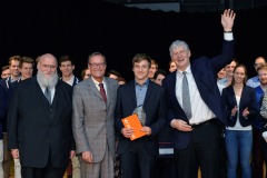 Mehrere Männer und Frauen stehen nebeneinander; ein Mann rechts hebt die Hand, einer hält eine orange Mappe.