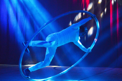 Person in weißer Kleidung balanciert in einem großen Metallreifen auf einer Bühne mit blauem Licht.