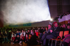 Publikum in einem Saal, oben große Licht- oder Nebelwolke, rechts sitzende Zuschauer in Reihen.