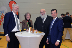 Vier Personen stehen an einem Stehtisch mit Gläsern, einer Flasche, Snacks und Unterlagen in einer Halle.