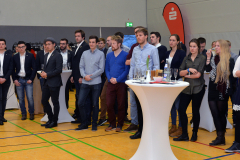 Personengruppe in einer Sporthalle, stehend an Stehtischen mit Getränken; Basketballkorb und rote Fahne im Hintergrund.