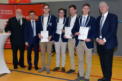 Sechs Männer stehen in einer Sporthalle, vier halten Urkunden; rote Banner und ein Stehtisch links.