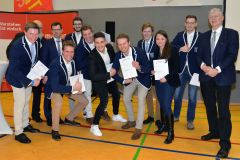 Gruppe von Männern und einer Frau in blauen Blazern, einige mit Urkunden, vor rotem Banner in einer Halle.