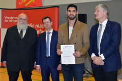 Vier Männer in Anzügen stehen vor einer roten Wand; einer hält ein Zertifikat.