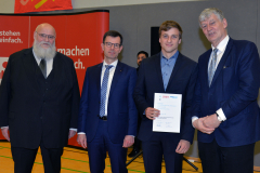 Vier Männer in Anzügen stehen vor einer roten Wand; einer hält eine Urkunde.