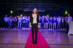 Frau steht auf rotem Teppich in einer Halle, dahinter eine Gruppe von Menschen; rechts ein Mann am Stehpult.
