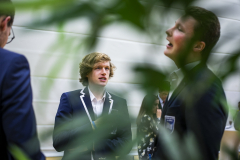 Drei Personen in dunklen Blazern stehen in einem hellen Raum, im Vordergrund unscharfe grüne Blätter.