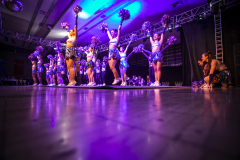 Cheerleader-Gruppe auf einer Bühne mit erhobenen Pompons unter violettem Licht; eine Person kniet rechts.