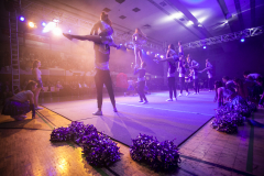 Cheerleader-Gruppe bei einer Show in einer Halle, mit erhobenen Posen und lila Pompons auf dem Boden.