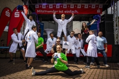 Gruppe von Menschen in weißen Kitteln und Sportkleidung posiert auf einer Bühne mit dem Schriftzug „Miteinander ist einfach.“
