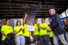 Gruppe von Menschen in leuchtend gelben T-Shirts auf einer Bühne, eine Person hält ein Schild, daneben steht ein Mann im Anzug mit Mikrofon.