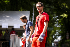 Mann in rotem T-Shirt und Handschuhen steht neben Mann in blauem T-Shirt vor einem Zelt im Freien.