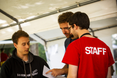 Drei Personen unter einem Zelt; eine trägt ein rotes T-Shirt mit der Aufschrift „STAFF“.