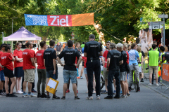Menschen in roten und schwarzen Shirts stehen auf einer Straße unter einem Ziel-Banner; rechts Absperrgitter und ein WC-Schild.