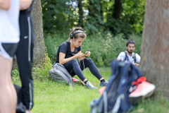 Person sitzt auf einer Wiese am Baum und schaut auf ein Smartphone; im Vordergrund sind Rucksäcke zu sehen.