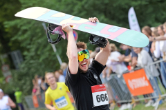 Person mit Startnummer 666 hält ein Snowboard über dem Kopf; im Hintergrund stehen Zuschauer hinter Absperrgittern.