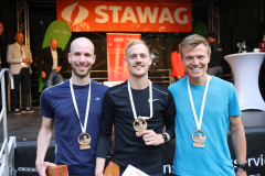 Drei Männer mit Medaillen stehen vor einer Bühne mit STAWAG-Banner; im Hintergrund stehen weitere Personen.