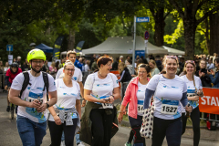 Läuferinnen und Läufer mit Startnummern gehen auf einer Straße; im Hintergrund Zelte, Bäume und Zuschauer.