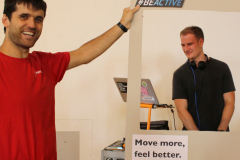 Zwei Männer, einer im roten Shirt hält einen Rahmen; Schild mit „#BEACTIVE“ und „Move more, feel better.“