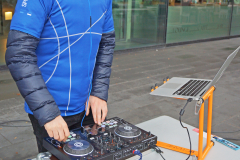 Mann in blauem T-Shirt steht an einem DJ-Mischpult neben einem Laptop vor einem Glasgebäude.