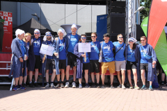 Gruppe von Menschen in blauen Shirts vor einer Bühne, einige halten Schilder; rechts Banner, links rotes Plakat.