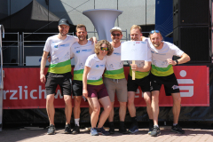 Sechs Personen in weißen T-Shirts stehen vor einer Bühne; eine hält ein Schild mit „CVT“.
