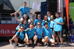 Gruppe von Personen in blauen T-Shirts posiert vor einer Bühne; eine Person sitzt auf den Schultern einer anderen.