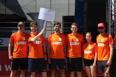 Sechs Personen in orangefarbenen Sportshirts stehen nebeneinander; eine hält ein Schild mit der Aufschrift „Roadrunners“.