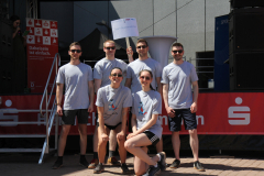 Fünf Personen vor einer Bühne, eine hält ein Schild „Team Supramolche“, rote und blaue Banner im Hintergrund.
