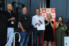 Fünf Personen stehen auf einer Bühne; zwei halten Papiere, eine Person klatscht, ein rotes Schild im Hintergrund.