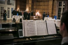 Piano mit Notenblatt im Vordergrund; im Hintergrund stehen mehrere Personen in einem holzgetäfelten Saal an Stehtischen.
