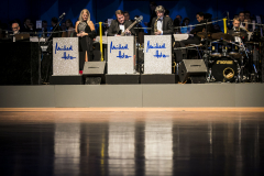 Orchester auf Bühne mit Notenpulten, Sängerin, Gitarrist, Saxophonist und Schlagzeug.