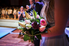 Frau hält einen Blumenstrauß mit Lilien und rosa Blüten; im Hintergrund stehen mehrere Personen.