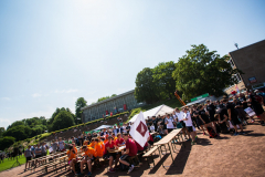 Viele Menschen sitzen und stehen auf einem Sportplatz vor Bänken, Zelten und einem Gebäude.