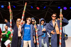 Mehrere Personen auf einer Bühne, einige im blauen Bademantel, halten lange, mit Klebeband umwickelte Stäbe hoch.
