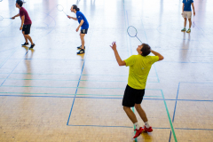 Mehrere Personen spielen Badminton in einer Sporthalle mit hohen Fenstern.